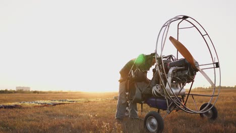 The-motor-paraglider-stands-in-the-field-at-sunset-with-a-wooden-propeller,-and-the-pilot-lays-out-the-parachute-and-aligns-the-slings.-flight-preparation-to-configure-the-fastening-of-the-helmets