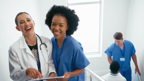 Portrait-Of-Female-Doctor-And-Nurse-With-Clipboard-Discussing-Patient-Notes-On-Stairs-In-Hospital