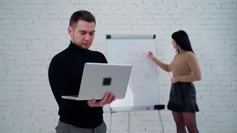 Young man and woman in the studio. Handsome man working on a laptop while woman is writing on a white board behind him.