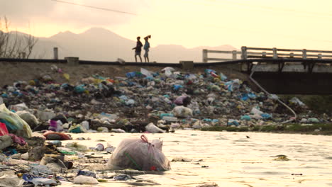 Silhouette-of-people-walking-over-bridge-with-piles-of-rubbish-on-side-slope-in-Vietnam