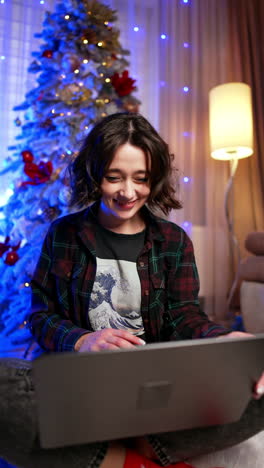 caucasian woman with short dark curly hair sits on the floor near christmas tree. portrait of a happy lady working online on laptop from home. vertical video.