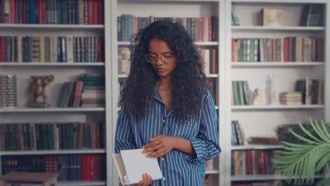 smart indian girl university student standing next to bookshelf holding textbook