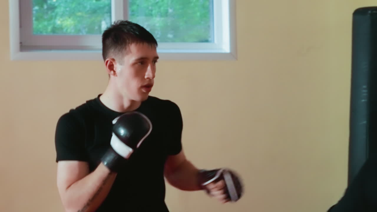 Pugilist wearing black shirt bends forward inside gym with red mat and boxing equipment visible, demonstrating defensive movement, energy, focus, and readiness during intense combat training session