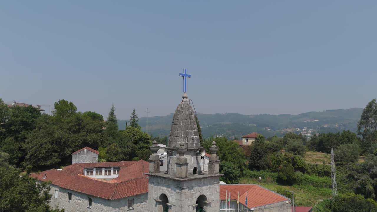 Aerial view of a granite church tower crowned by a vivid blue cross over the Minho hills