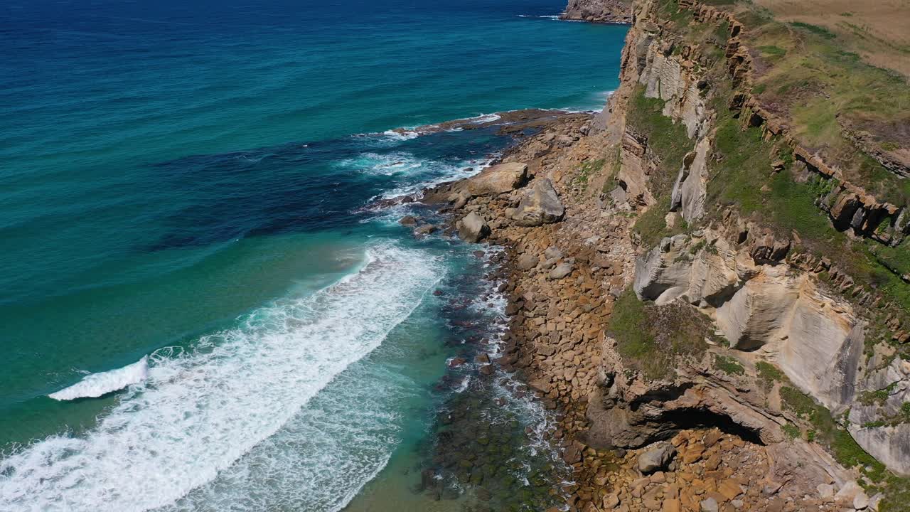 Lateral flight on a sunny summer day over a cliff with green grass and orange rocks that goes deep into the turquoise sea over the calm waves of white foam in the Cantabrian Sea Spain