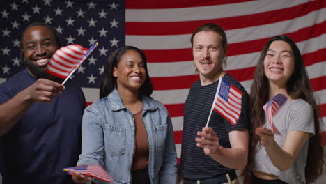 Studio-Portrait-Shot-Of-Multi-Cultural-Group-Of-Friends-Waving-Miniature-Stars-And-Stripes-Flags-In-Front-Of-American-Flag-Celebrating-4th-July-Independence-Day-Party-2