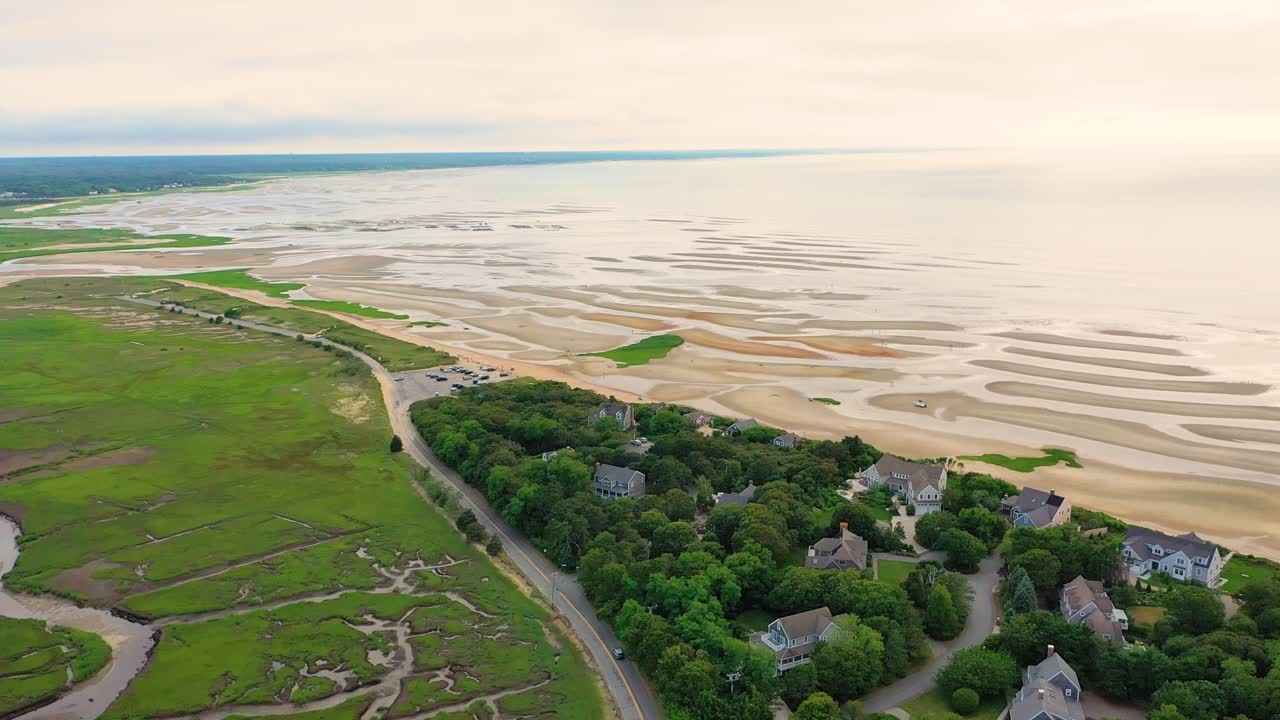 Cape Cod public beach at low tide reveals sculpted sand ridges, shimmering tide pools, and exposed flats, bordered by seaside houses, coastal marshland, and cars parked near the shoreline