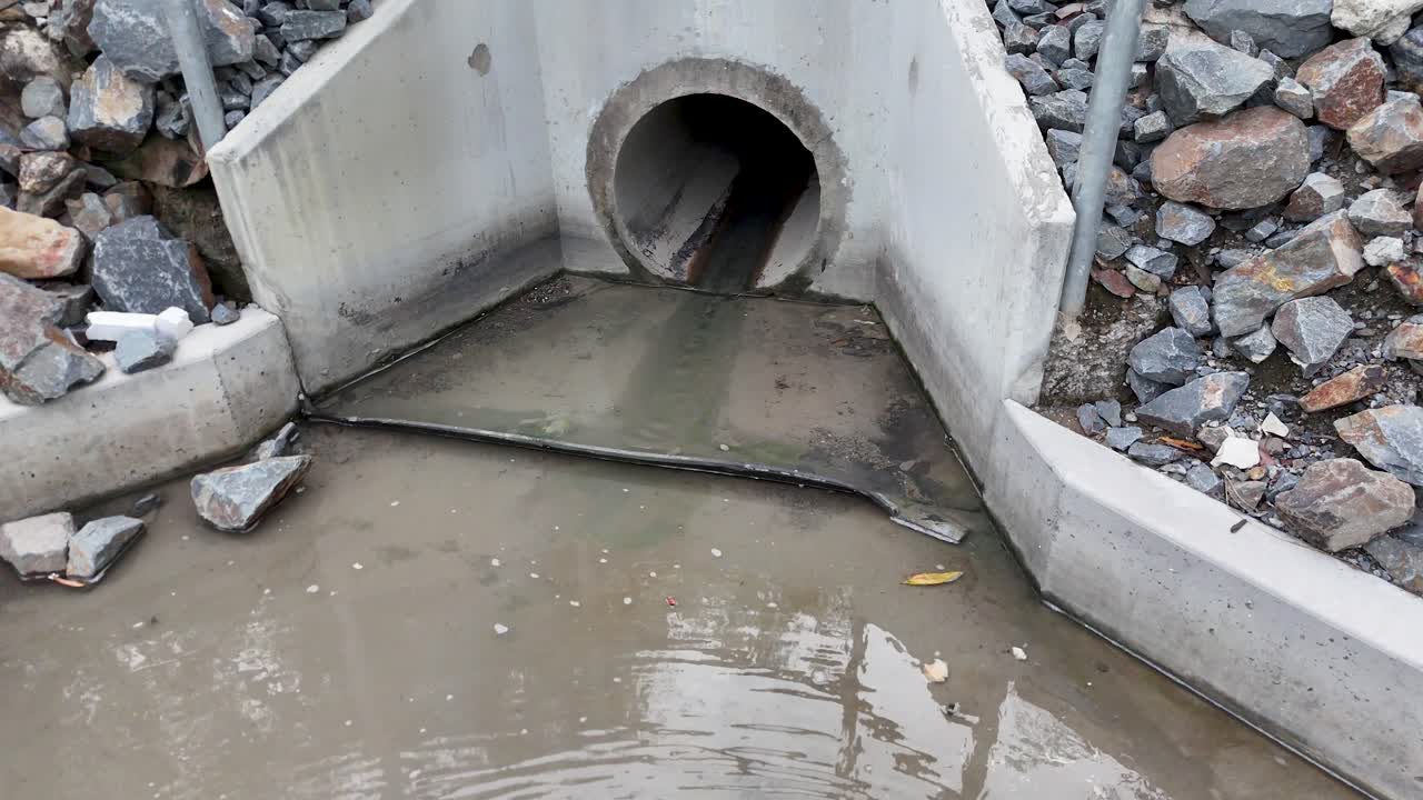 Steady stormwater flows from concrete culvert into rocky channel, overcast daylight, fixed camera view