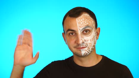 man with painted face is waving hand and looking at camera against blue background