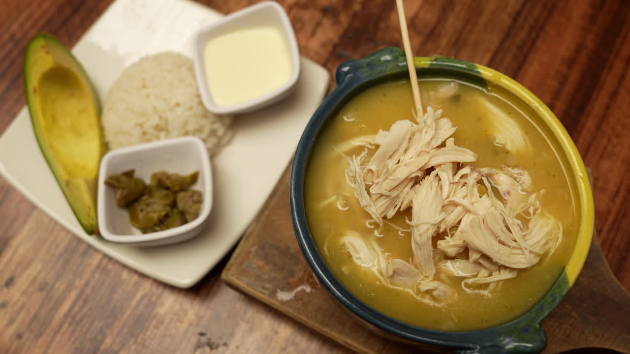 A warm, inviting bowl of Colombian ajiaco served with creamy avocado, hearty rice, zesty capers, and rich cream, set against a rustic wooden table for a homely touch