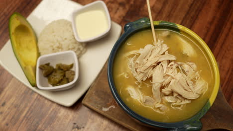 a warm, inviting bowl of colombian ajiaco served with creamy avocado, hearty rice, zesty capers, and rich cream, set against a rustic wooden table for a homely touch