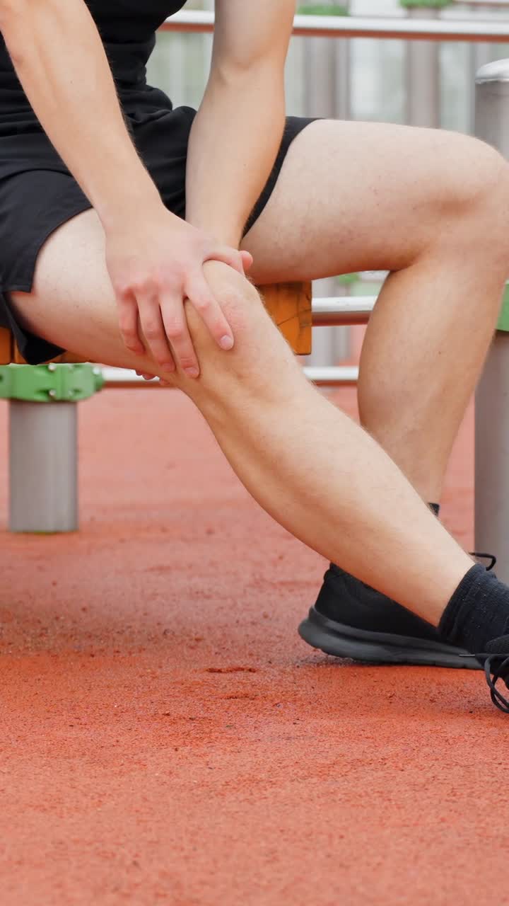 Injured caucasan athlete sitting down holding his knee, feeling a sharp pain after a workout, experiencing joint problem, health issue in his leg, vertical static slow motion close up