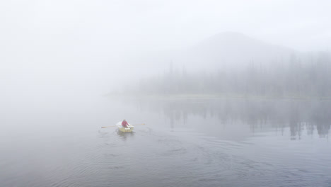 person rowing a boat in white, foggy lake landscape with mountain and forest in the background