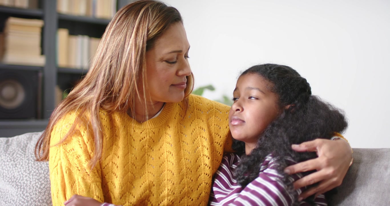 African American mother noticing and holding child in striped top withdrawing on sofa to calm