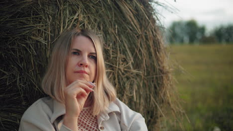 lady reflecting with something in her mouth, adjusting her head on hay bale in open field, peaceful rural setting with warm tones, dry grass, and scattered hay bales