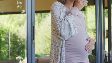 Mittelteil-Einer-Kaukasischen-Schwangeren-Frau,-Die-Mit-Einer-Tasse-Tee-Am-Fenster-Steht-Und-Den-Bauch-Berührt