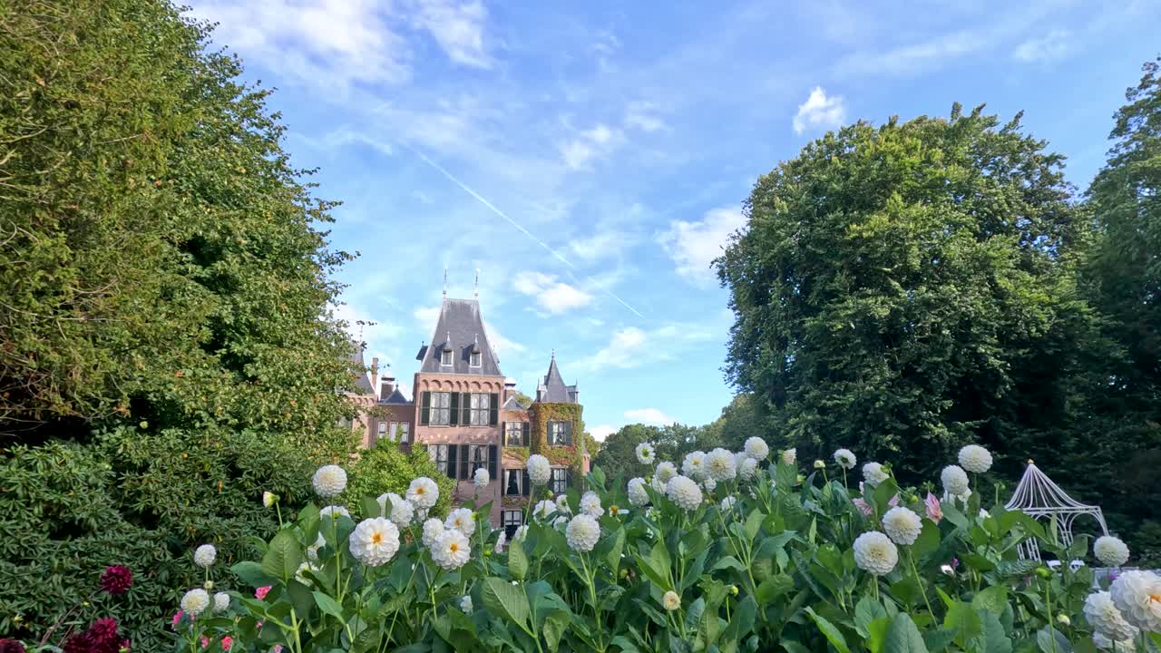 Camera glides through lush botanical garden with blooming dahlias, revealing a grand historic castle under bright daylight and clear blue sky