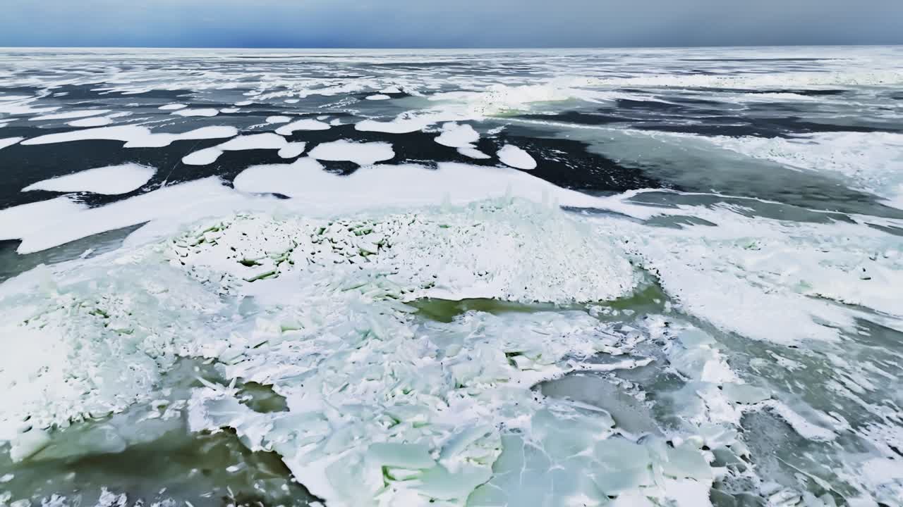 Планирование дрона с воздуха над растрескавшейся замерзшей водой со снежными пятнами, торосами и темными оттаявшими участками в драматическом зимнем морском пейзаже.