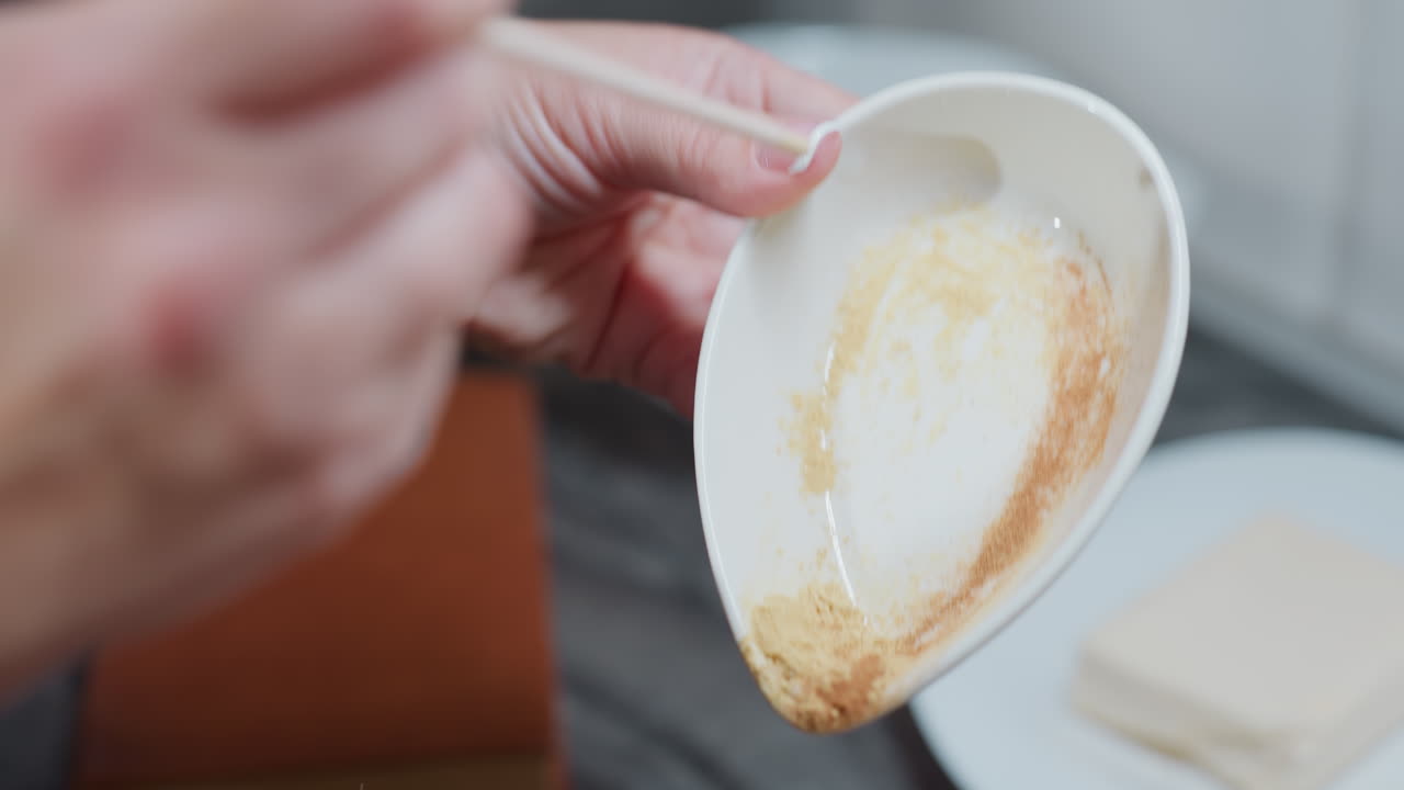 primer plano de la mano sosteniendo un plato blanco con ingrediente en polvo, raspando los restos con un palo antes de agregarlo a la olla, fondo borroso con la encimera y los ingredientes