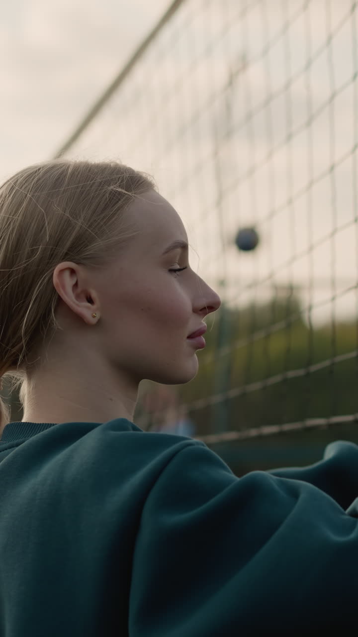 Close-up of young lady in green sweater checking firmness of volleyball net with blurred view of someone playing volleyball in the background during an outdoor sports training session