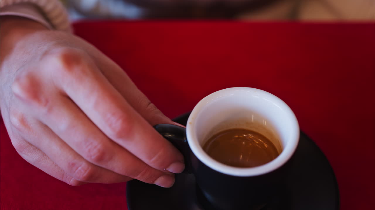 Close up of a woman holding an espresso in a black cup on a red tablecloth at a restaurant