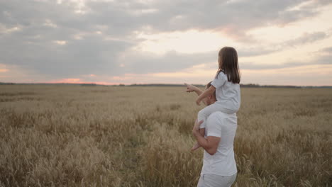 view from the back: daddy carries on his shoulders his beloved little healthy daughter in sun. in slow motion the daughter walks with her father on the field and free and happy waves her hands up.