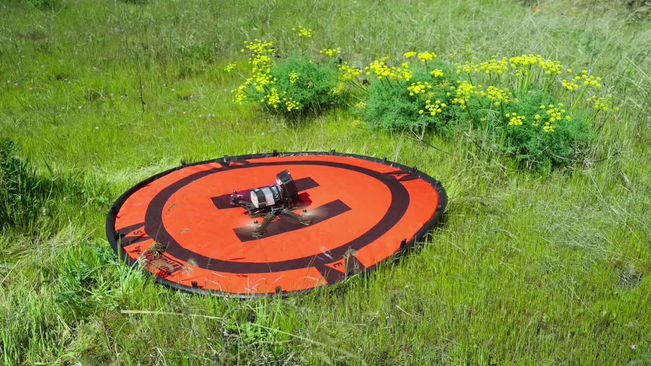 A First Person View (FPV) drone with a mounted GoPro action camera slowly taking off like a rocket from an orange hoodman helipad as the wind blows through green grass and yellow flowers