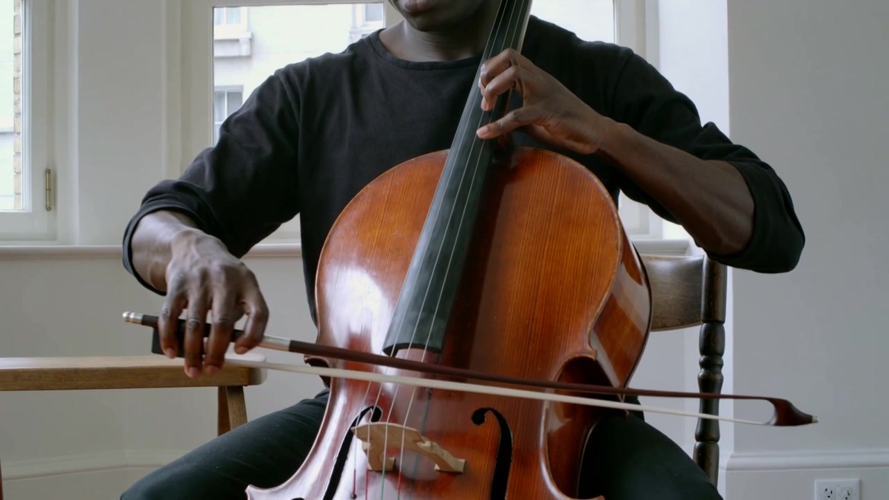 Starting male cellist drawing bow across cello strings at window, wearing black shirt, practicing