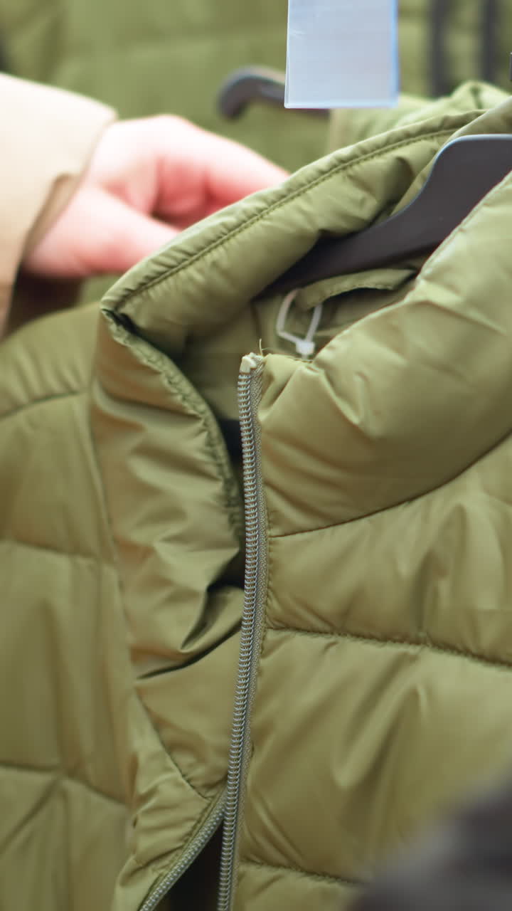 un primer plano de una mano con una chaqueta marrón alcanzando una chaqueta verde de un estante de ropa en un centro comercial. destaca la textura de la chaqueta hinchada y la naturaleza casual de las compras
