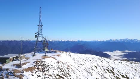 An-aerial-pass-over-a-snow-covered-New-Zealand-mountain,-highlighting-a-sturdy-communications-tower-amid-pristine-winter-scenery
