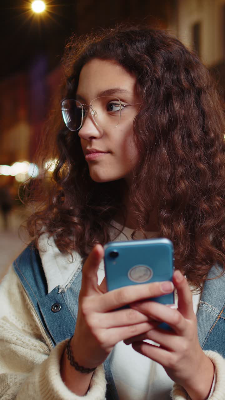 Happy smiling teenager girl in eyeglasses using smartphone typing text messages in urban city street