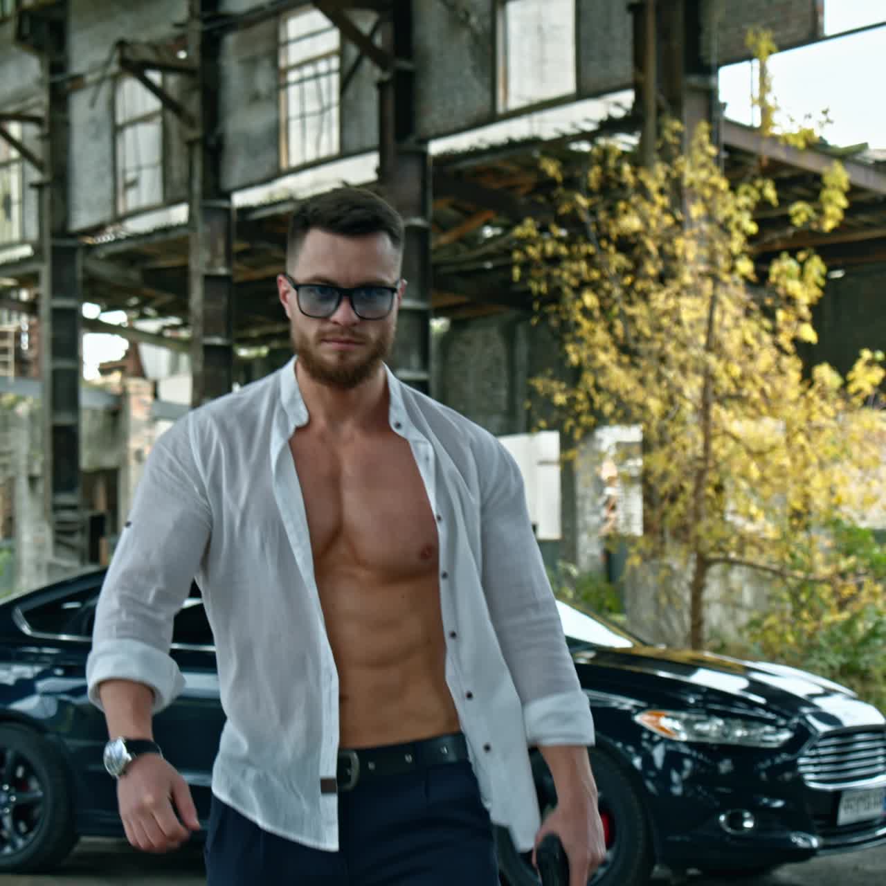 Handsome man with a weapon in abandoned place. Sporty guy in stylish glasses and white unbuttoned shirt with a pistol in his hand on black car background.
