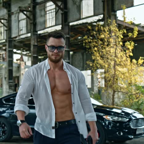 handsome man with a weapon in abandoned place. sporty guy in stylish glasses and white unbuttoned shirt with a pistol in his hand on black car background.