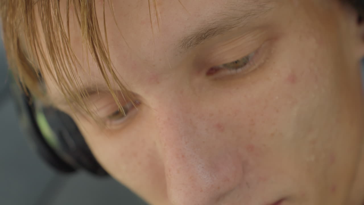 primo piano di uomo caucasico che ascolta musica con le cuffie, attore introspettivo che prova le battute all'aperto, cuffie over-ear, luce solare soffusa sulla pelle lentigginosa, sudore leggero, sguardo verso il basso, profondità di campo ridotta