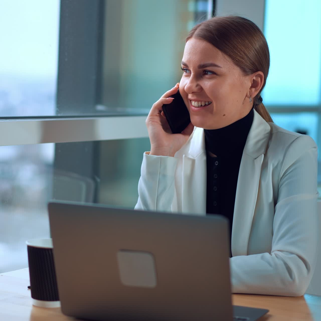 Business lady having phone conversation sitting with laptop in front of her. Woman talks and smiles looking at window