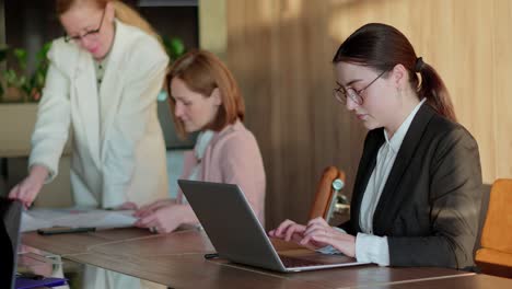 Nahaufnahme-Eines-Selbstbewussten-Brünetten-Mädchens-Mit-Runder-Brille-Und-Business-Kleidung,-Das-An-Einem-Laptop-Arbeitet-Und-Auf-Der-Tastatur-Tippt,-Während-Es-Zusammen-Mit-Seinen-Kollegen-Am-Tisch-Im-Büro-Arbeitet