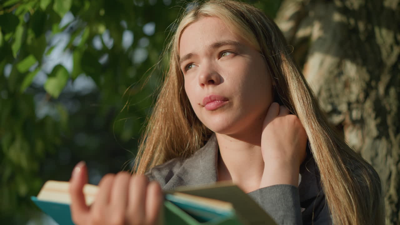 나무 에 앉아 있는 회색 옷을 입은 여인이 왼손으로 목을 만지며 사려 깊은 표정으로 먼 곳을 바라보며, 빛이 그녀의 얼굴을 부드럽게 비추고, 그녀는 열린 책을 들고 있다.