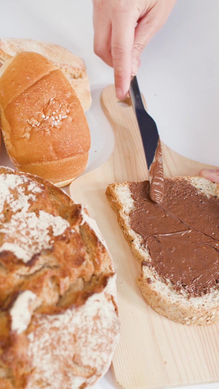 Close-up of hand spreading chocolate cream on sourdough bread amidst various fresh bakery assortments.