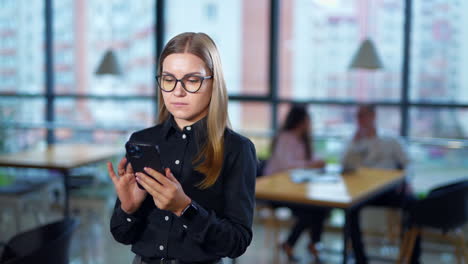 young woman in glasses texting somebody on smartphone and smiling. two office workers sitting at the table and talking at the background in blur.