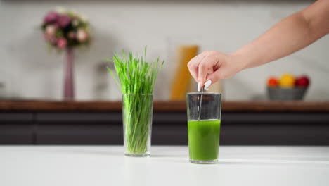 hand with white nails inserting a straw into a tall glass of green wheatgrass juice