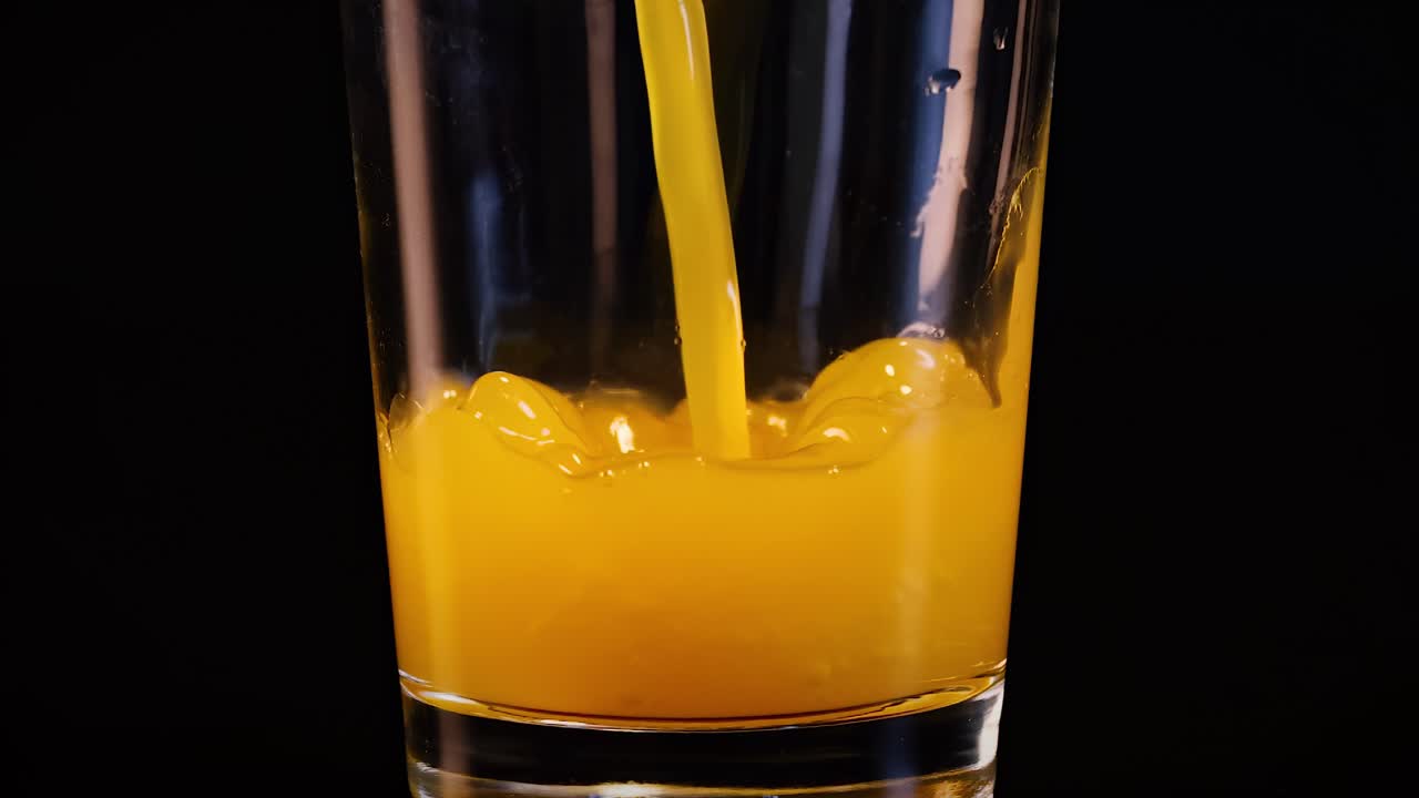 A bright yellow liquid is dynamically poured into a clear glass against a dark background.