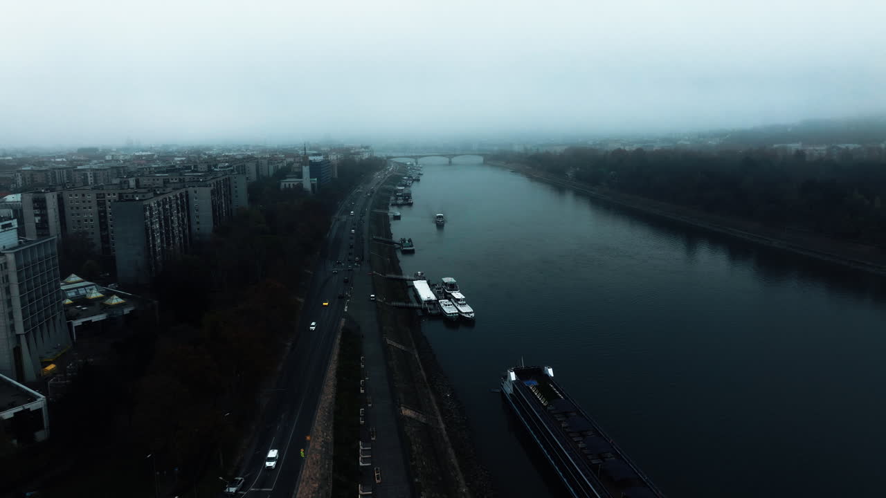 A dark, misty view of the Danube in Budapest on a cold, overcast day. Fog, smog, and distant residential blocks create a bleak Eastern European atmosphere with muted, moody tones