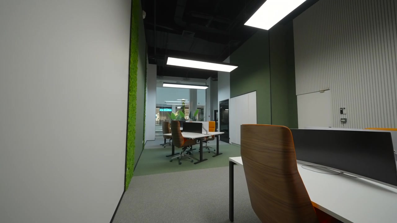 Narrow view into a contemporary office interior with a vertical green plant wall on the left, clean white walls, gray flooring, and desks and chairs visible