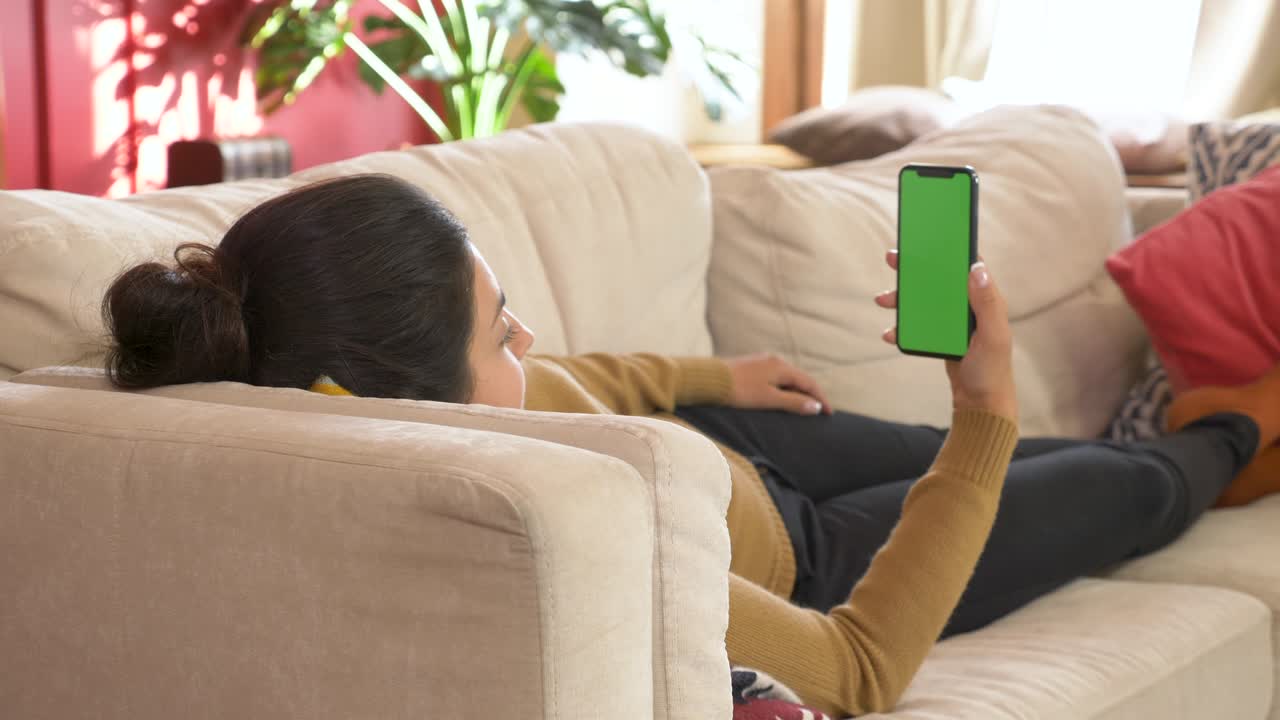 소파에 누워있는 젊은 인도 여성은 손에 빈 디지털 전화를 들고, 휴대 전화 녹색 화면, 수직 모드에서 모의 화면