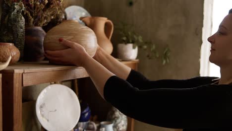 Side-view-portrait-of-contemporary-female-potter-putting-handmade-vase-on-shelf-in-studio,-pleased-with-her-work,-smiling-to-the-camera.-Slow-motion