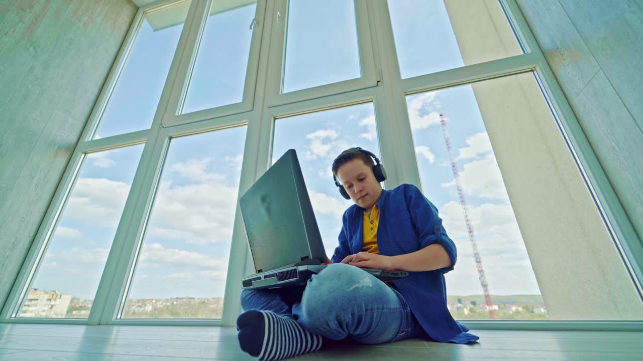 Gaming at home. Boy playing video games in his leisure time. Teenager in earphones sitting near the window and playing on a laptop. Happy boy wins the game.