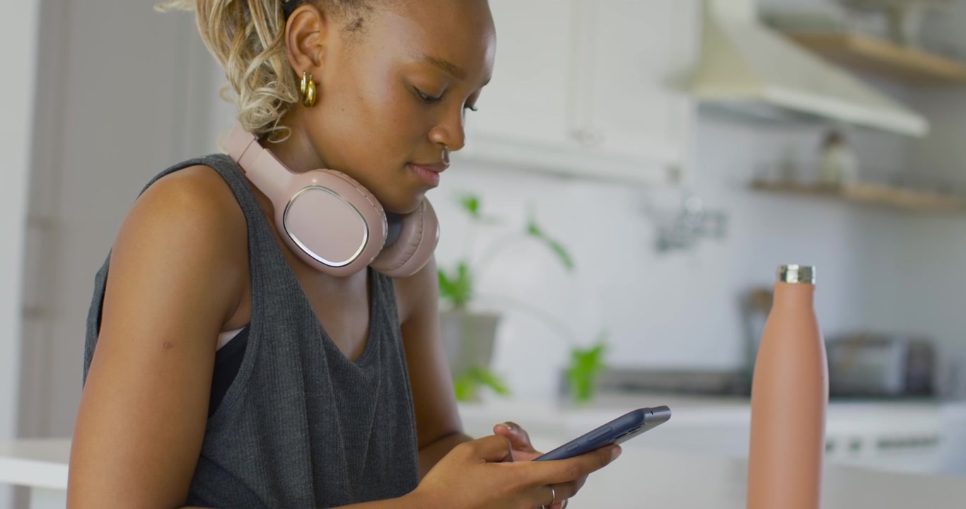 African woman glancing, picking up, scrolling smartphone at kitchen counter with pink headset