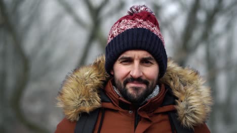 bearded caucasian male in warm clothes looks at camera and smiles in the forest during cold winter day, slow motion