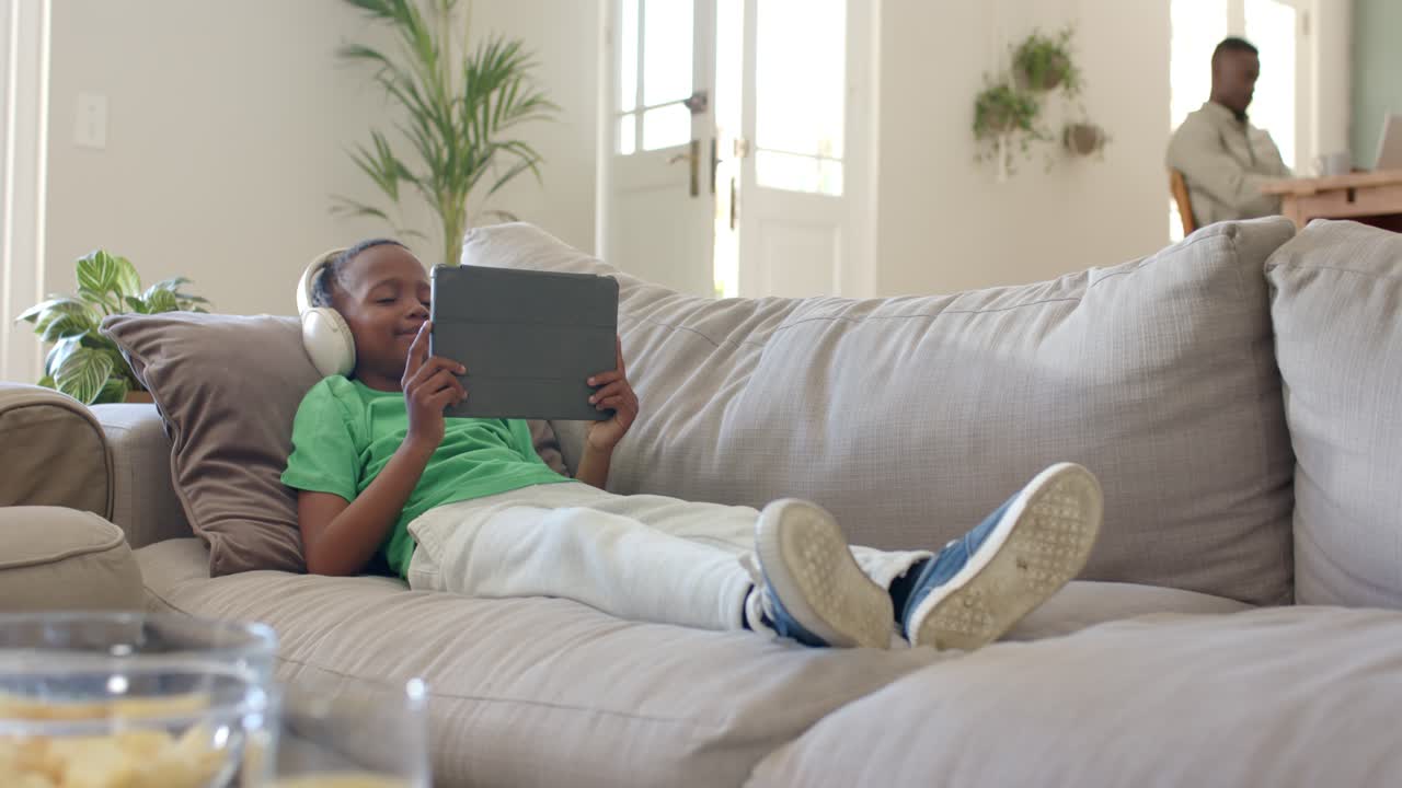 African American father and son lying on sofa at home with headset watching tablet father working