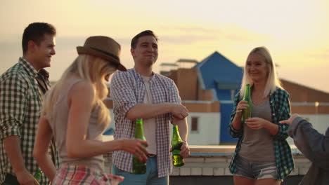 a company of six young people smile and communicates on the roof and drink beer on a summer evening. two girls are dancing at this moment.